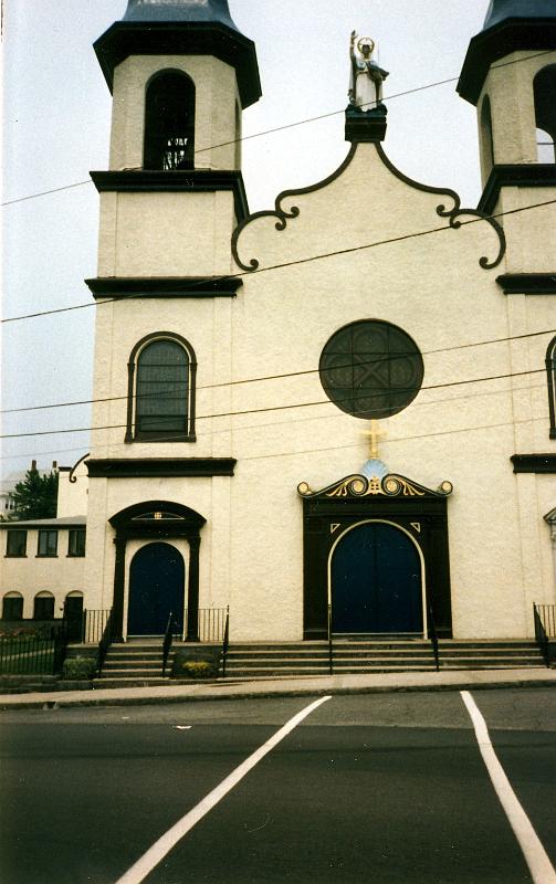 01 Originally dedicated in 1893, Our Lady of Good Voyage was built for the Portuguese community in Gloucester, after they petitioned the Roman Catholic Church for the establishment of a place to worship dedicated to the Madonna. Large numbers of Portuguese immigrants migrated from the rugged Azores Islands and began settling around Gloucester's Inner Harbor as early as 1829 to work in the city's active fishing industry. By 1888, approximately 200 Portuguese families lived in Gloucester making it the largest Portuguese colony on the East Coast. According to the story of Our Lady of Good Voyage, a stranded fisherman in the rough Atlantic Ocean broke one of his oars and could not return to his homeport. He sought help from the Madonna and the sea miraculously calmed allowing him to reach port safely. A fire destroyed the original church in 1914. Prominent architect Halfdan M. Hanson designed and immediately began building the existing, unique Mission style church, which replaced the earlier church. It is the only Mission style church in Gloucester. Modeled after a church in the Azores, Our Lady of Good Voyage consists of two distinct sections: the two-story main worship space that is of a cruciform plan and an L-shaped rectory that extends from the northwest corner of the main worship space. The rectory, which was built between 1872 and 1884 as a separate building, was incorporated into the new church. Resting on a granite foundation, the building is covered in a buff-colored stucco. Flanked by two identical bell towers, the central bay of the façade is pierced by the main entrance at the first level. A rose window adorns the second level, above which rises an ogee pediment supporting a pedestal and a statue of Our Lady of Good Voyage, who holds a boat in her left hand as a symbol of a safe voyage. In 1922, bells were installed in the towers. These bells, still in place today, were cast by John Taylor & Company of England-the same foundry that cast Phildelphia's Liberty Bell.